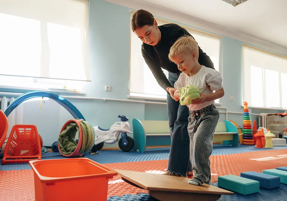 Mozgásfejlesztő Stephens-Sarlós Mozgásterápia-foglalkozáson részt vevő gyermek, aki egy egyensúlyozó táblán lépdel a mozgásterapeuta segítségével